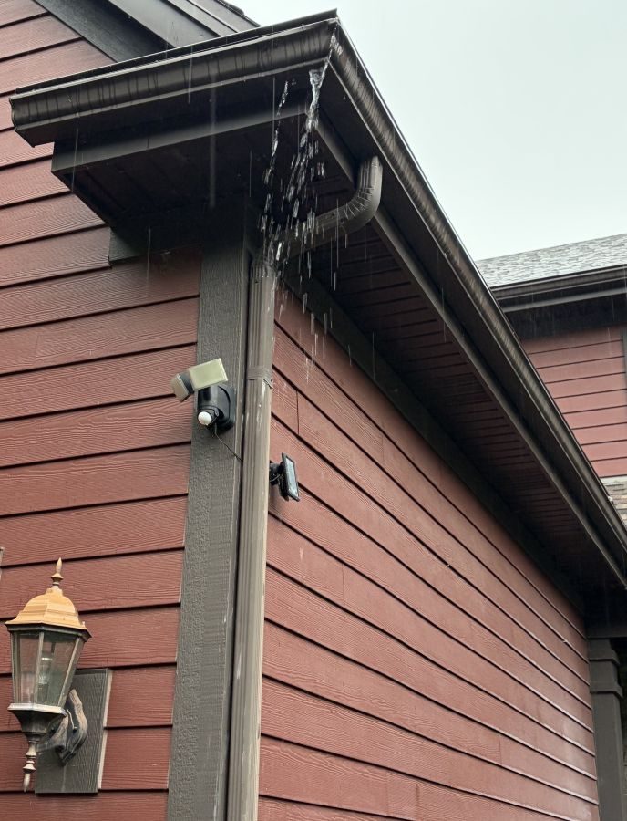 Rainwater overflowing the front edge of a gutter beside a downspout during a storm